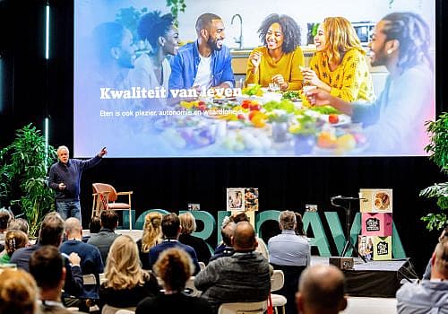 Spreker presenteert voor een zaal met bezoekers, met een groot presentatiescherm op de achtergrond