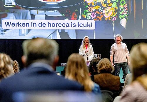 Twee sprekers staan op een podium voor een zaal met bezoekers tijdens een symposium, op het scherm staat 'werken in de horeca is leuk'
