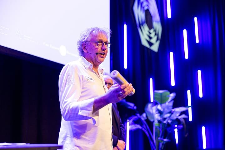 Jef Schuur spreekt op het podium en toont een object aan het publiek, met blauw-paars licht op de achtergrond