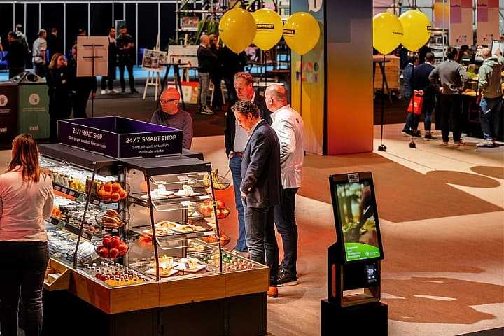 Bezoekers bekijken een vitrine met etenswaren bij een 24/7 Smart Shop op de beursvloer van Horecava, met displays, ballonnen en stands op de achtergrond