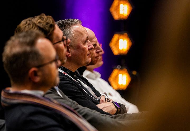 Rij mannen in het publiek luistert aandachtig tijdens een evenement, met sfeerverlichting op de achtergrond