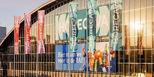 Voorzijde van RAI Amsterdam met grote Horecava-vlaggen en een banner in het zonlicht.