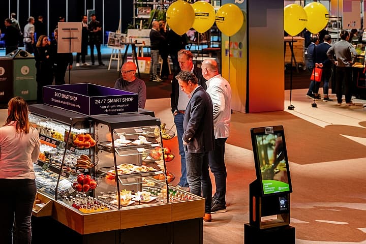 Bezoekers bekijken een vitrine met etenswaren bij een 24/7 Smart Shop op de beursvloer van Horecava, met displays, ballonnen en stands op de achtergrond