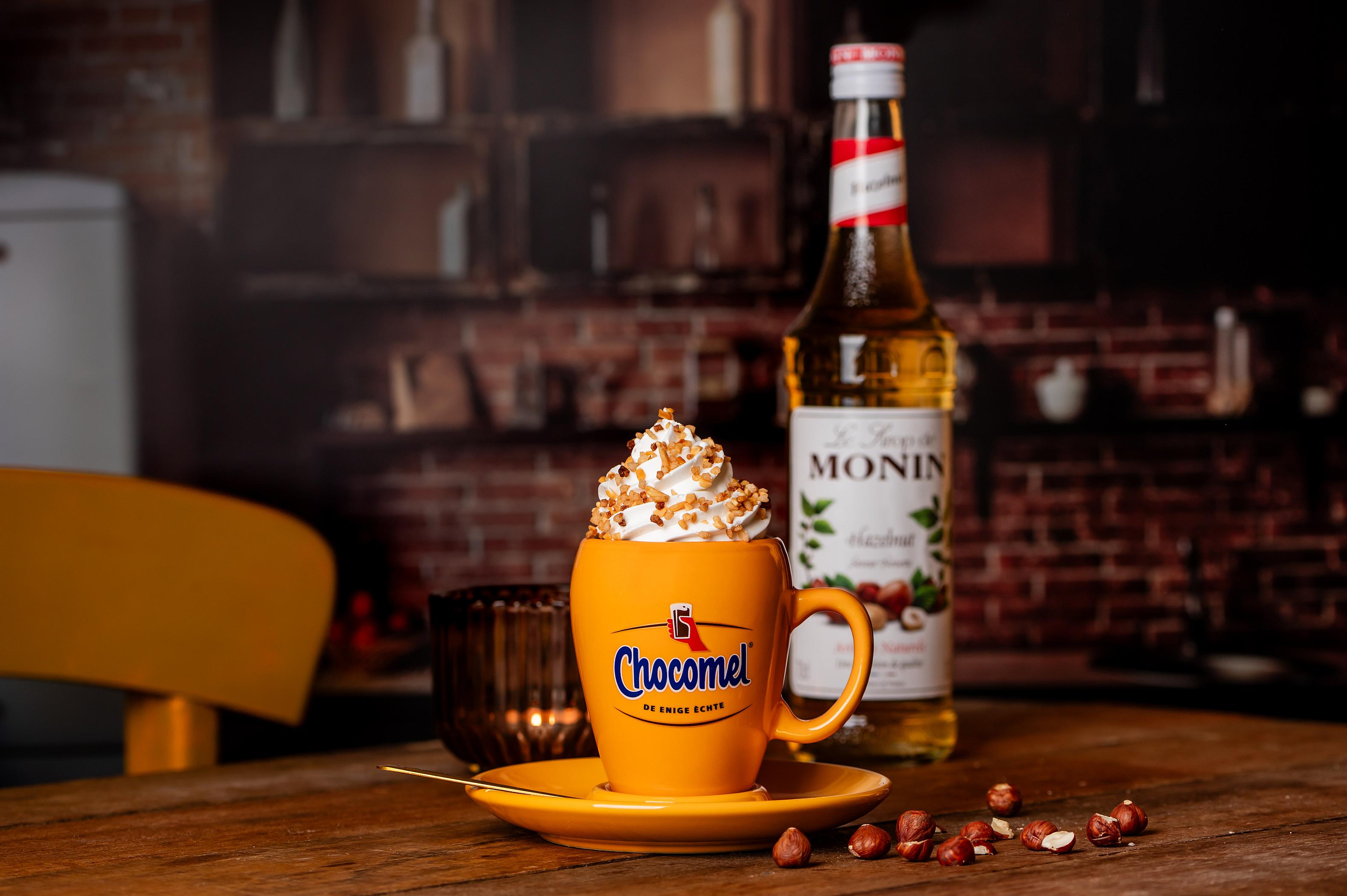 Een gele Chocomel-mok met warme chocolademelk, slagroom en gehakte hazelnoten, naast een fles Monin Hazelnoot siroop