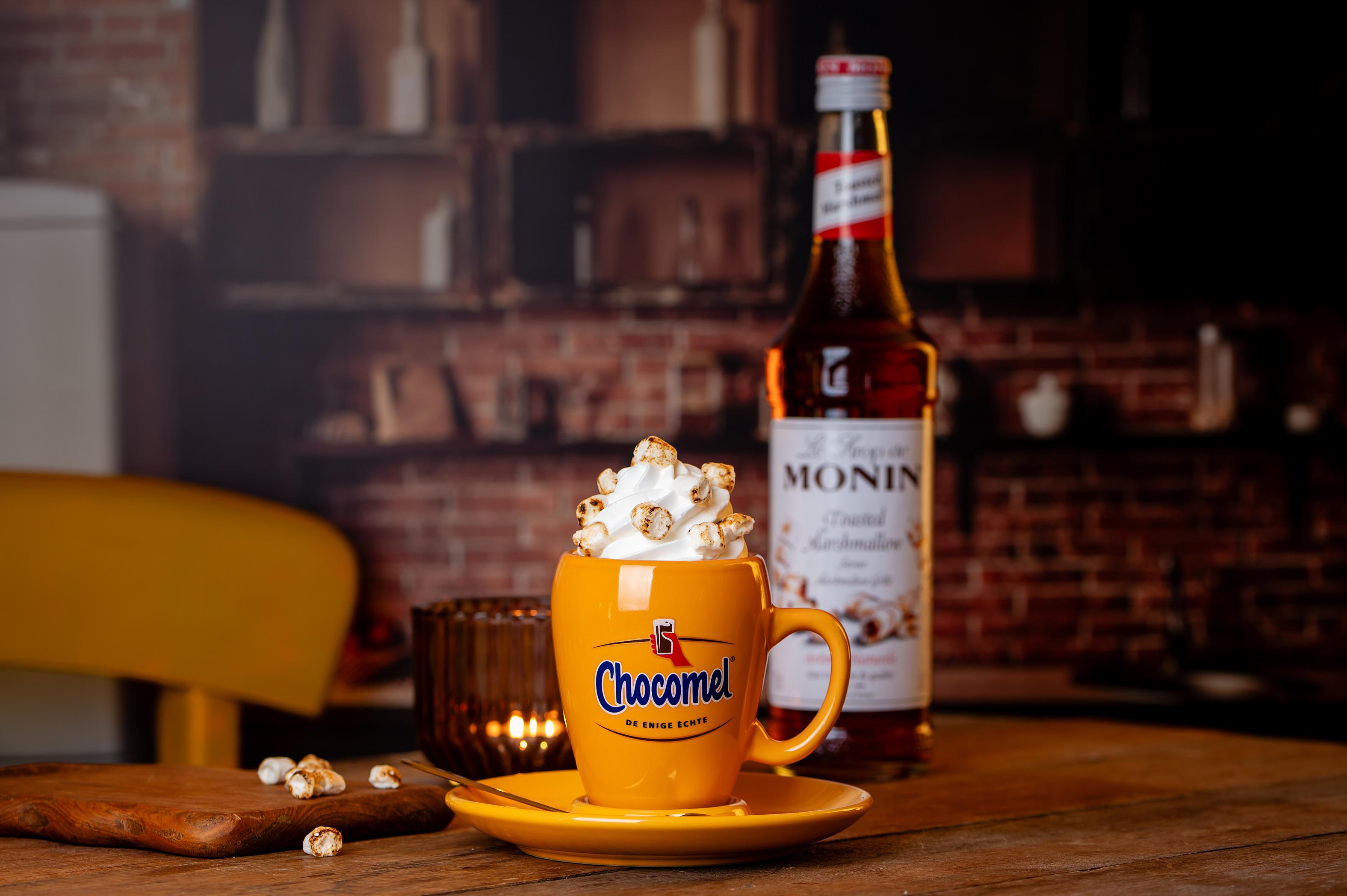 Een gele Chocomel-mok met warme chocolademelk, slagroom en geroosterde marshmallows, naast een fles Monin Toasted Marshmallow siroop